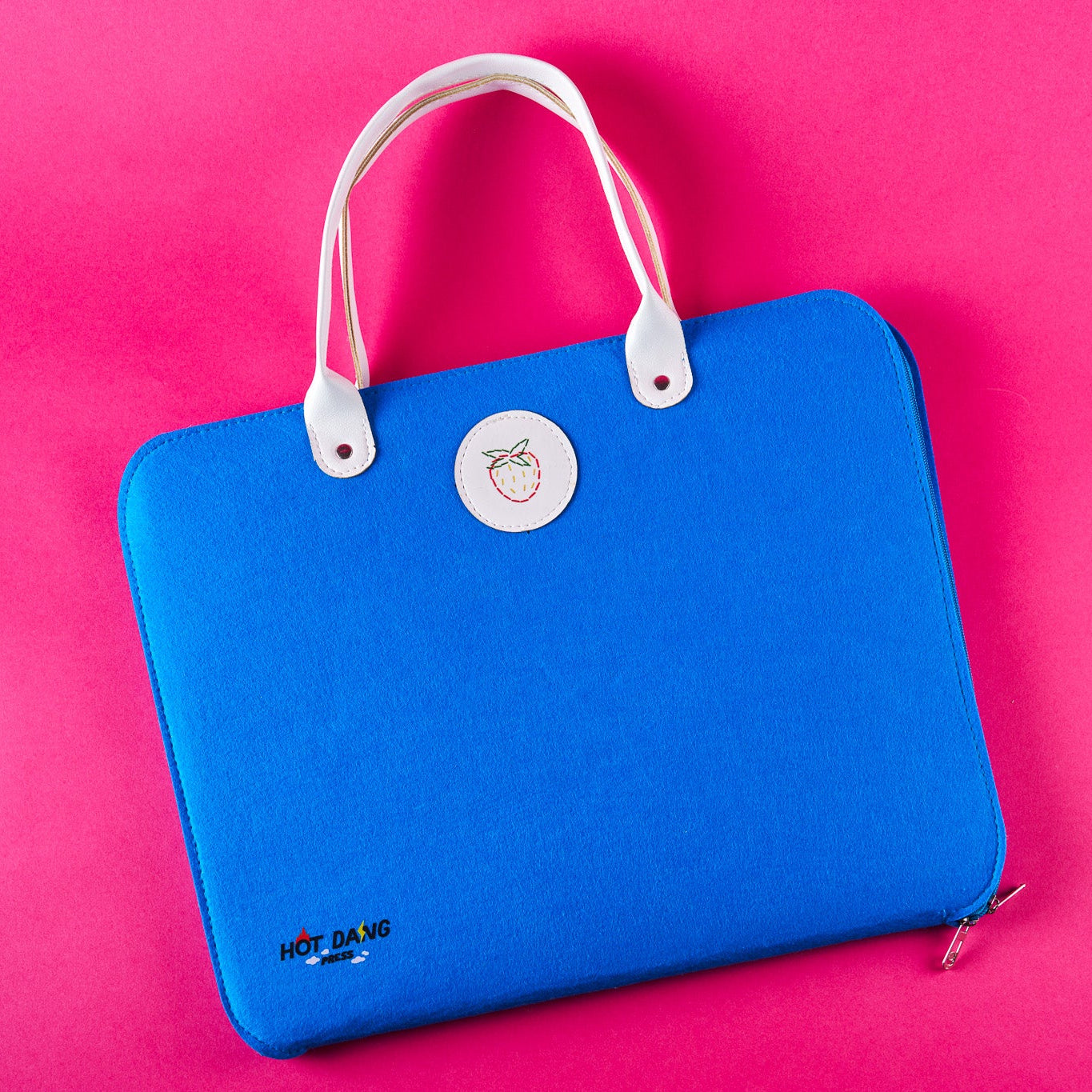 A bright blue felt carrying case with white top handles and a white circular logo patch, photographed on a solid hot pink background.