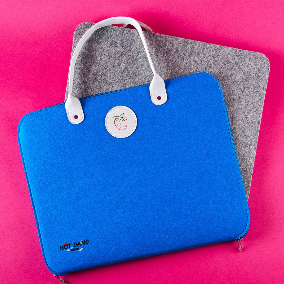 A bright blue felt sleeve with white handles and a circular white logo patch, shown on a pink background with a gray felt ironing mat underneath.