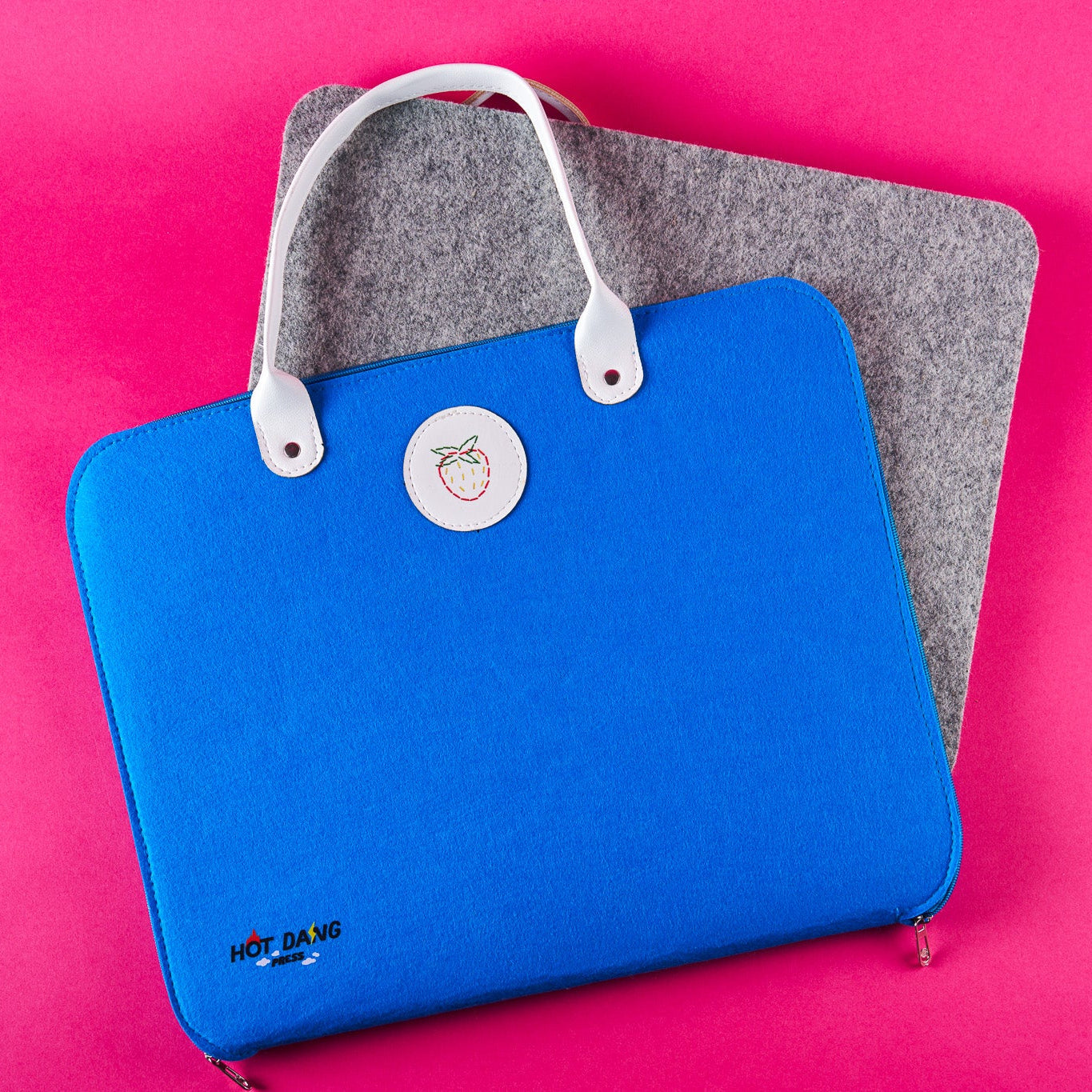 A bright blue felt sleeve with white handles and a circular white logo patch, shown on a pink background with a gray felt ironing mat underneath.