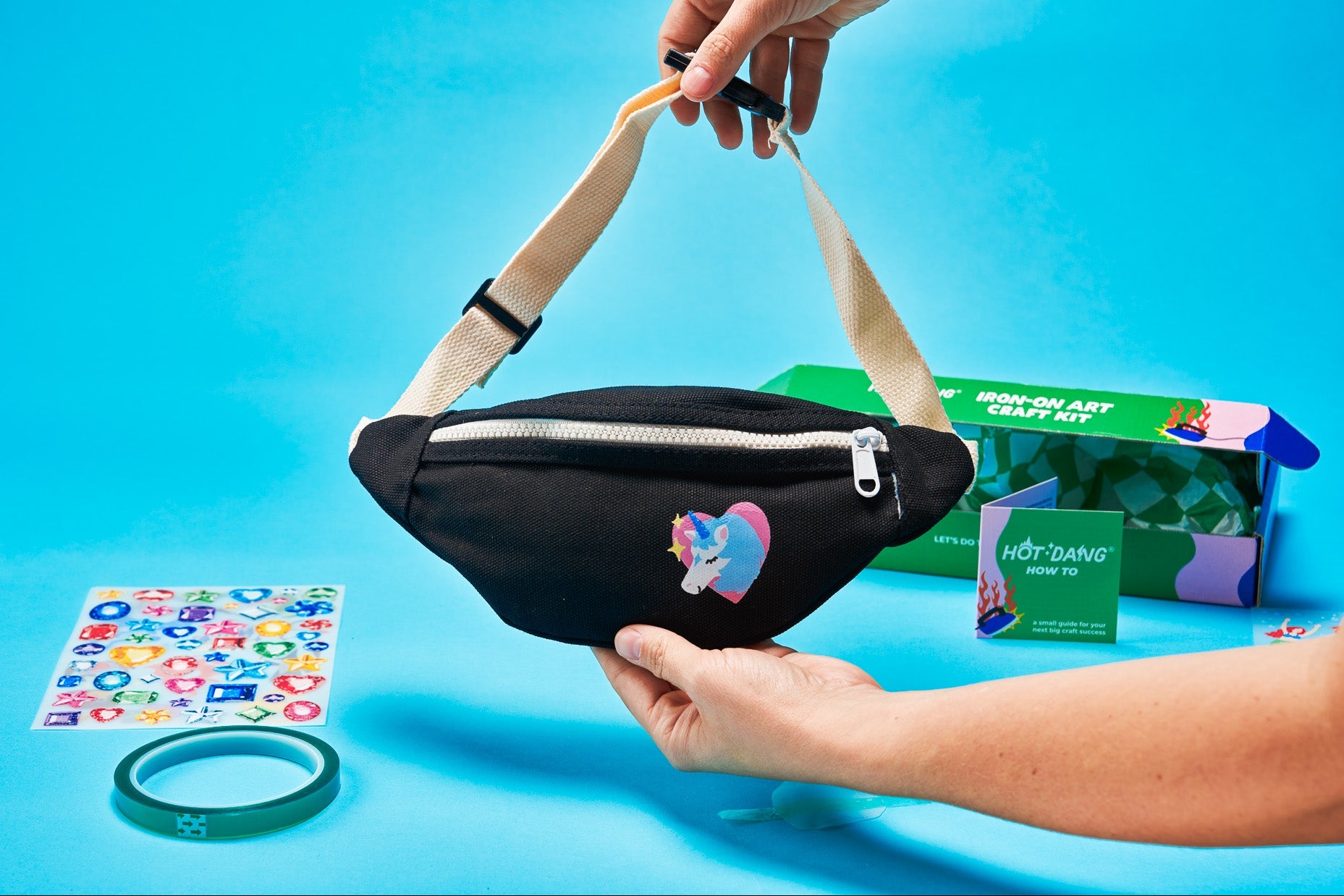 A black fanny pack with a beige strap is held by two hands against a bright blue background. The pack features a colorful unicorn iron-on transfer.