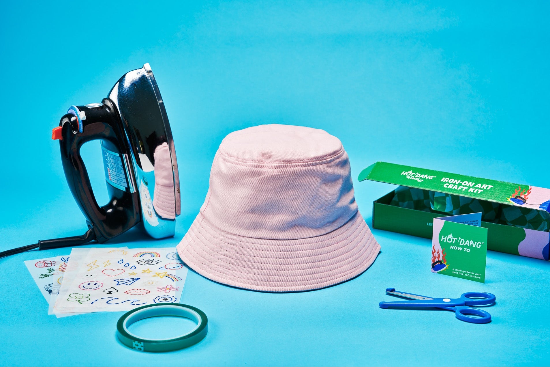 A pink bucket hat displayed with a black and silver iron, a green dtf craft kit box, blue-handled safety scissors, a green tape roll, and transfer sheets on a bright blue background.