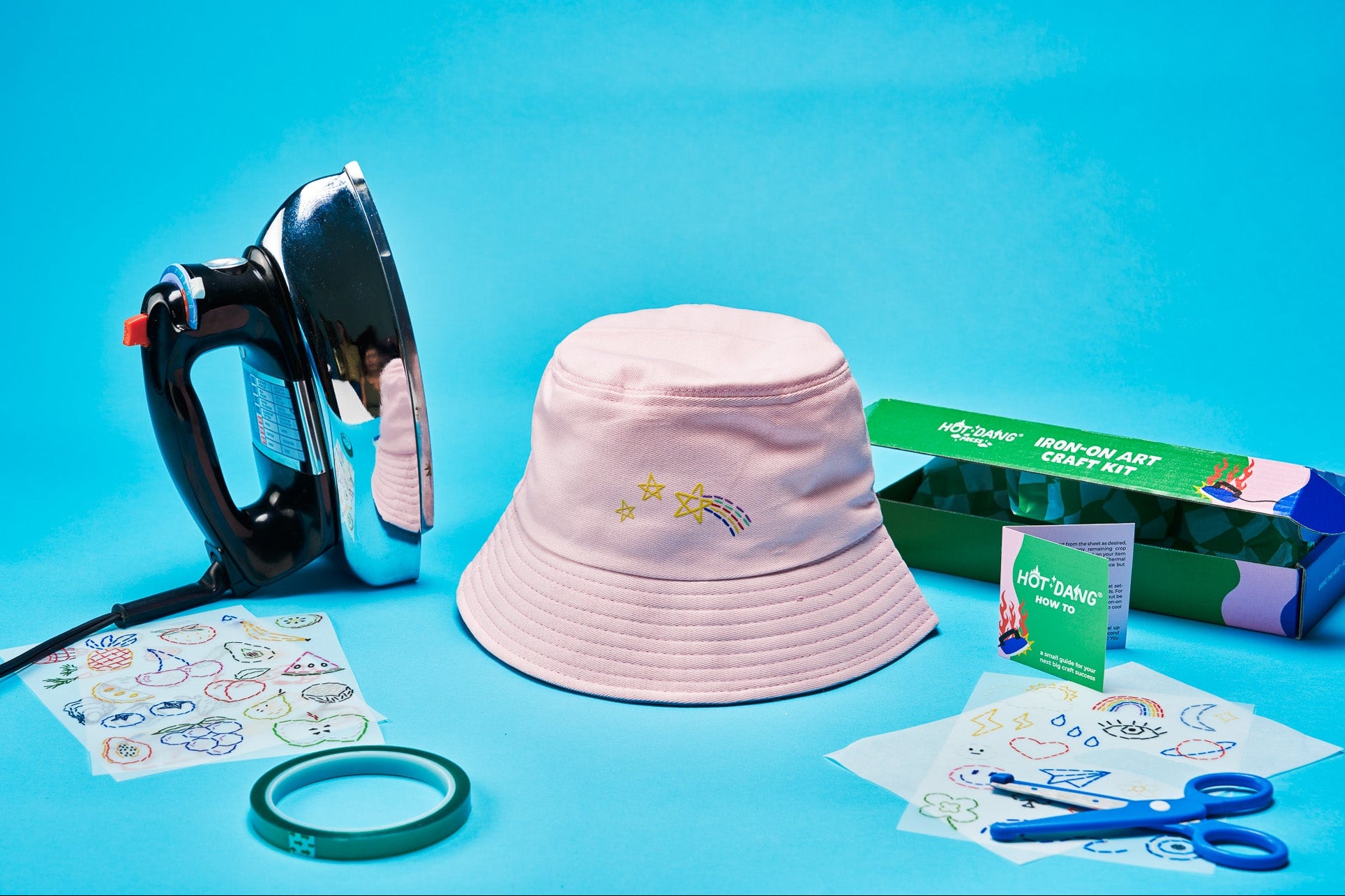 A pink bucket hat with iron-on embroidered stars and a shooting star lies on a bright blue background, surrounded by a black and silver iron, a green heat-resistant tape roll, dtf transfer sheets, blue-handled scissors, and a green 'Iron-on Art Craft Kit' box.